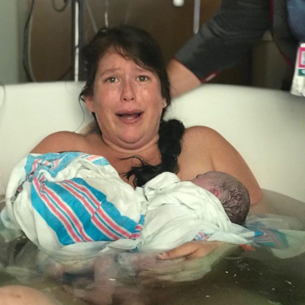 A woman in a birthing tub looks shocked while holding two newborn babies wrapped in blankets.