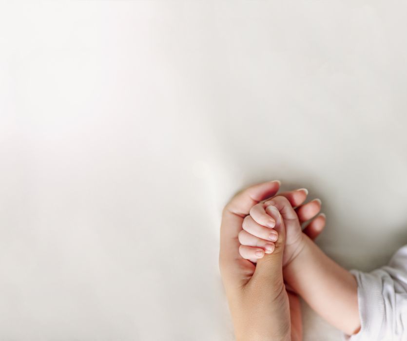 Close-up of a woman's hand gently cradling a baby's small fist on a soft white background.