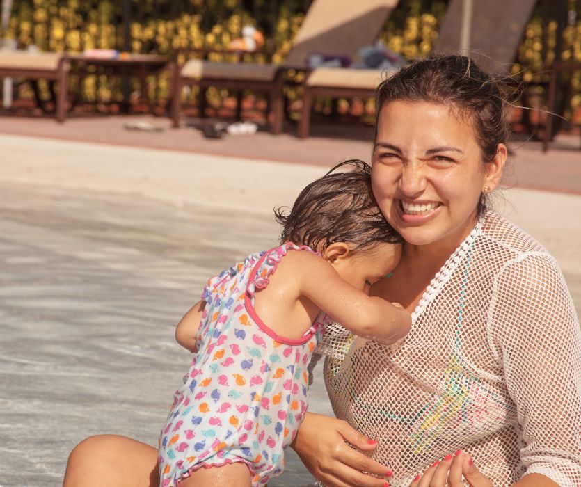 Smiling woman in a white mesh cover-up hugging a toddler in a patterned swimsuit at a shallow pool.