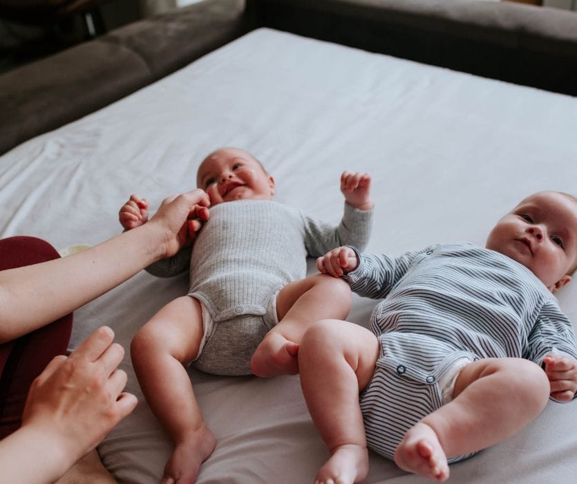 Twin babies lying on a white bed while a woman reaches out to touch one of them.