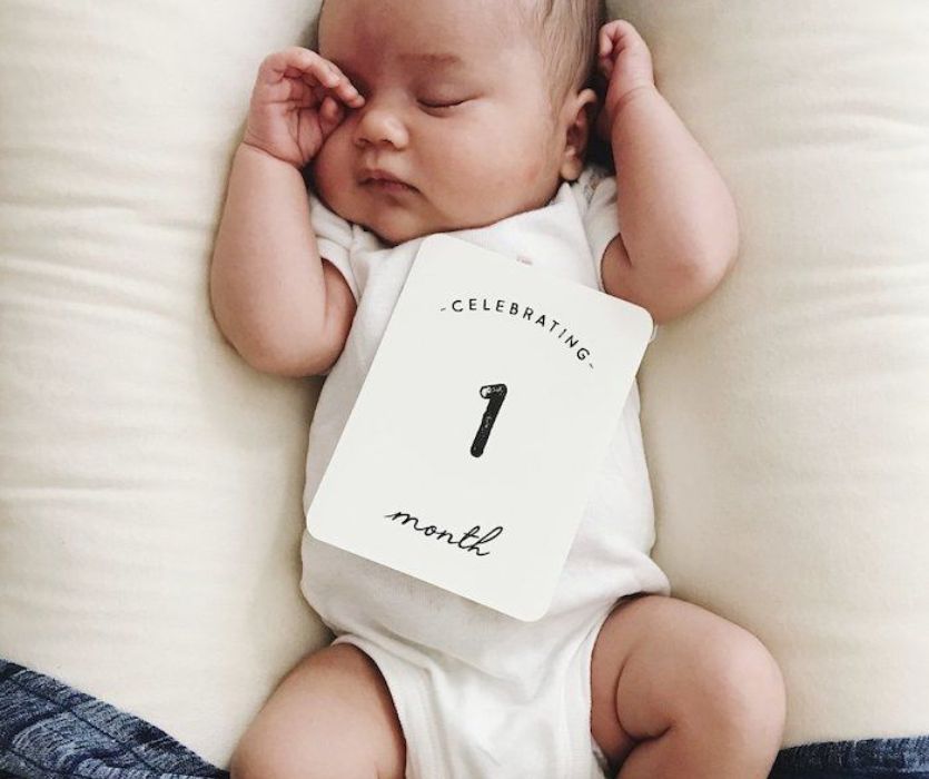 Sleeping infant laying on a white pillow with a milestone card that reads, Celebrating 1 month.