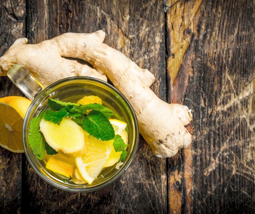 Ginger tea with lemon slices and mint leaves in a glass mug next to a fresh ginger root and lemon on a wooden table.