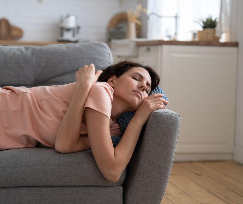 A woman lying on a gray sofa with her eyes closed, appearing unwell or tired.