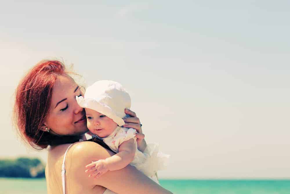 A red-haired mother holding a baby in a white sun hat at the beach.