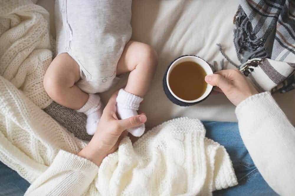 A person holding a cup of tea and a baby's foot while sitting on a bed among knitted blankets.