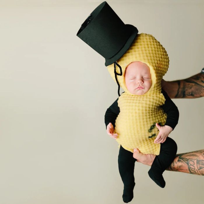 An infant wearing a Mr. Peanut costume with a textured yellow shell and a black top hat, being held by tattooed arms.