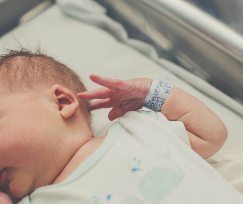 A close-up of a newborn baby lying in a hospital bassinet, showing their ear and arm with an identification wristband.