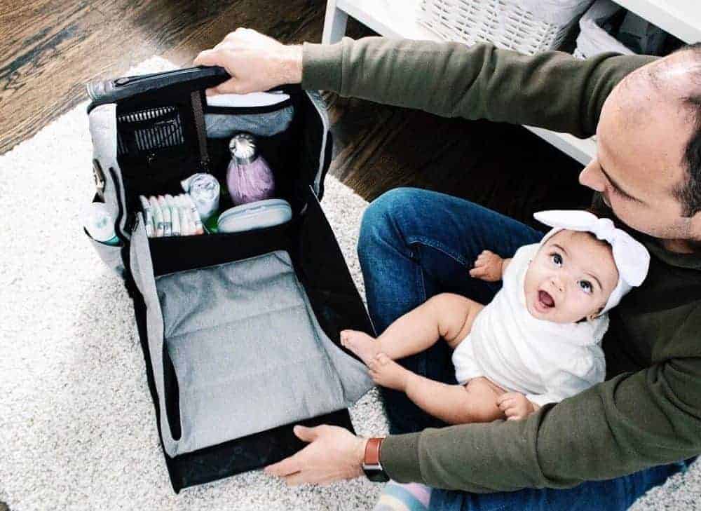 A man holds a baby while opening a light gray diaper bag backpack with a built-in fold-out changing station.