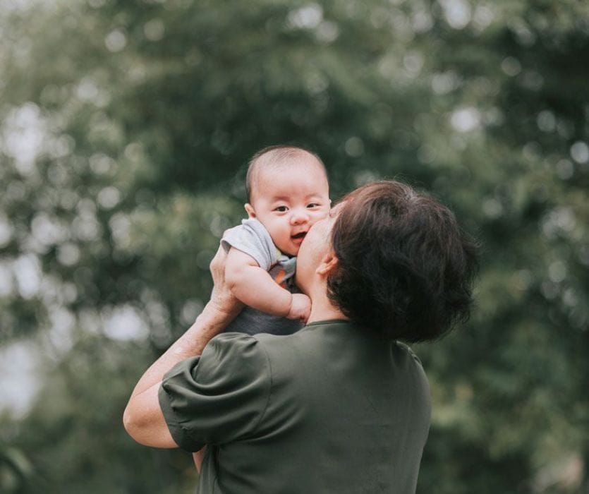 An older woman kisses the cheek of a baby she is holding up outdoors against a blurred green background.