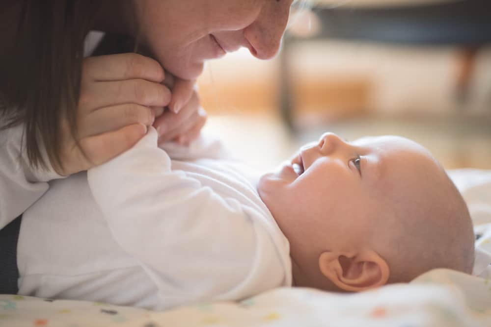 A smiling infant lying on their back looks up at a woman leaning over them.