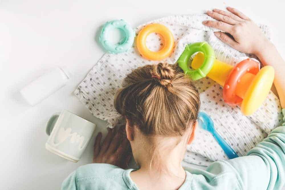 A woman rests her head on a table surrounded by baby toys, a bottle, a spoon, and a mug.