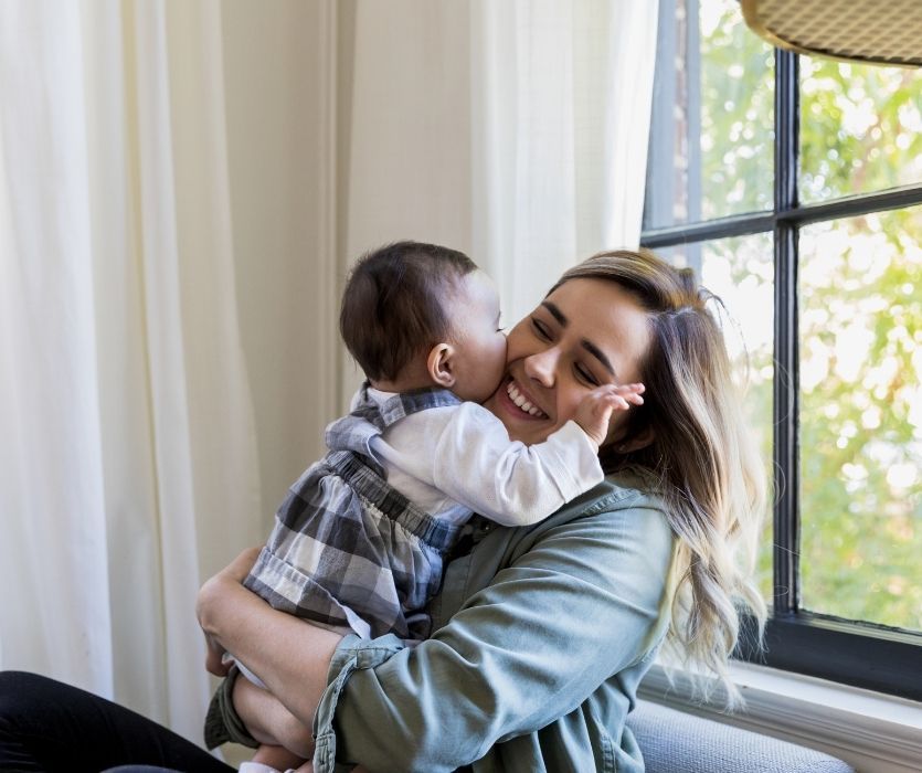 A smiling woman sitting by a window, hugging a baby who is kissing her on the cheek.