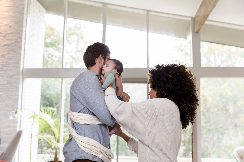 Parents standing in a bright living room, carefully adjusting their infant in a white fabric baby wrap carrier.