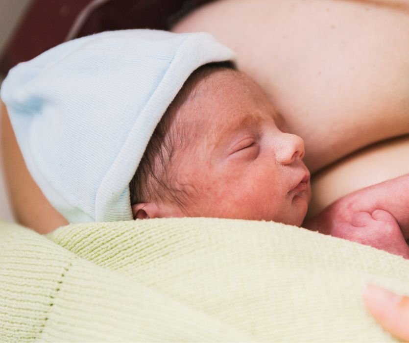 A sleeping newborn wearing a blue hat, wrapped in a lime green blanket during skin-to-skin contact with a parent.