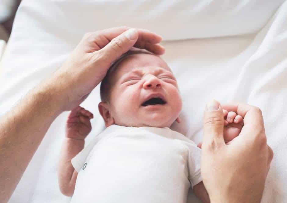 Crying newborn baby being comforted by an adult's hands on their head and hand.