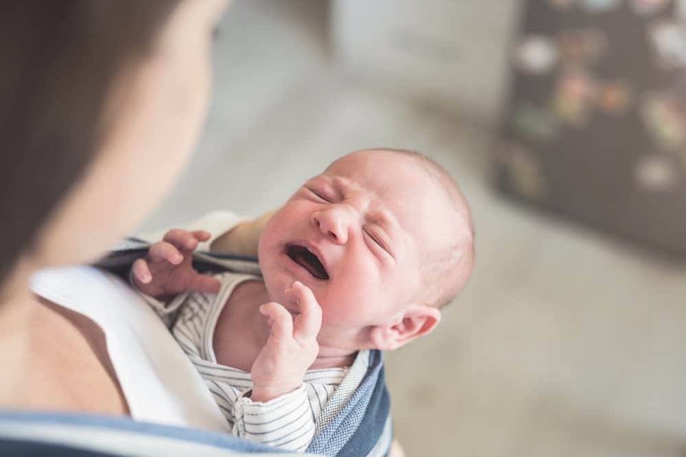 A crying newborn baby with eyes squeezed shut, being held in a fabric baby carrier by a parent.