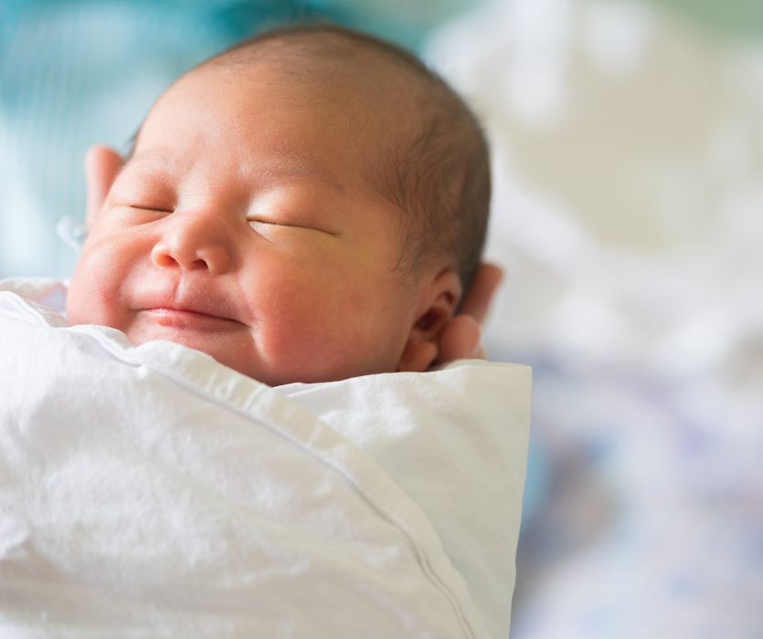 A smiling newborn baby swaddled in a white blanket.