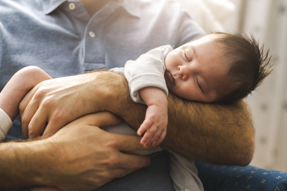 Newborn baby with dark hair sleeping peacefully while being cradled in a man's arms.