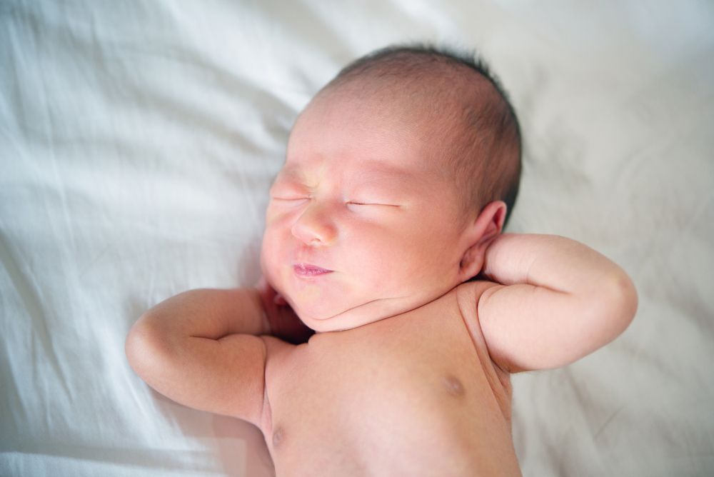 Close-up of a shirtless newborn baby sleeping on a white sheet with hands tucked behind the head.