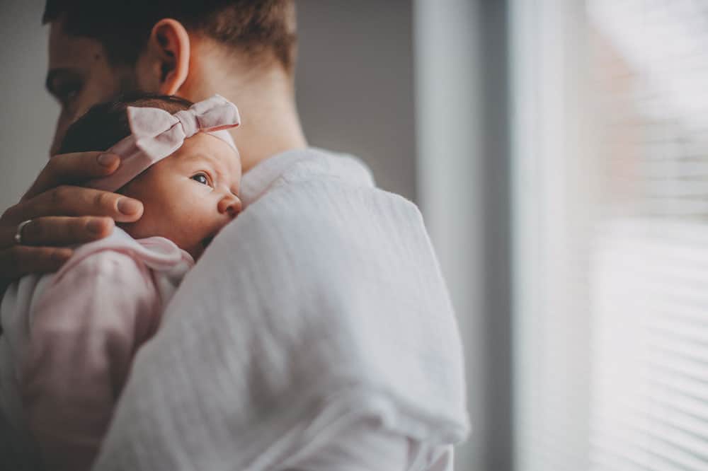 A father holds a newborn baby girl wearing a pink bow headband over his shoulder.