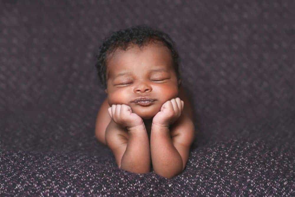 A sleeping newborn with his chin resting on both fists lies on a dark textured blanket, echoing the early newborn phase discussed in the post.