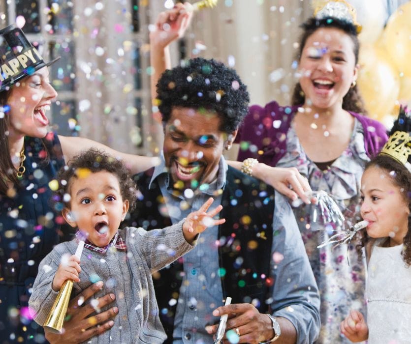 A diverse group of adults and children laughing and celebrating as colorful confetti falls around them.