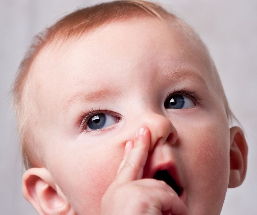 Close-up of a baby looking upward while pressing a finger against their nose.