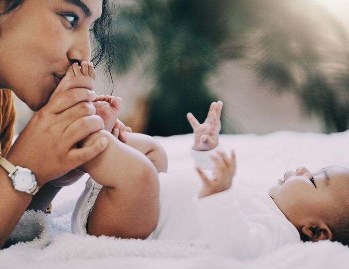 A mother kisses her baby's feet as the infant lies on a white blanket.