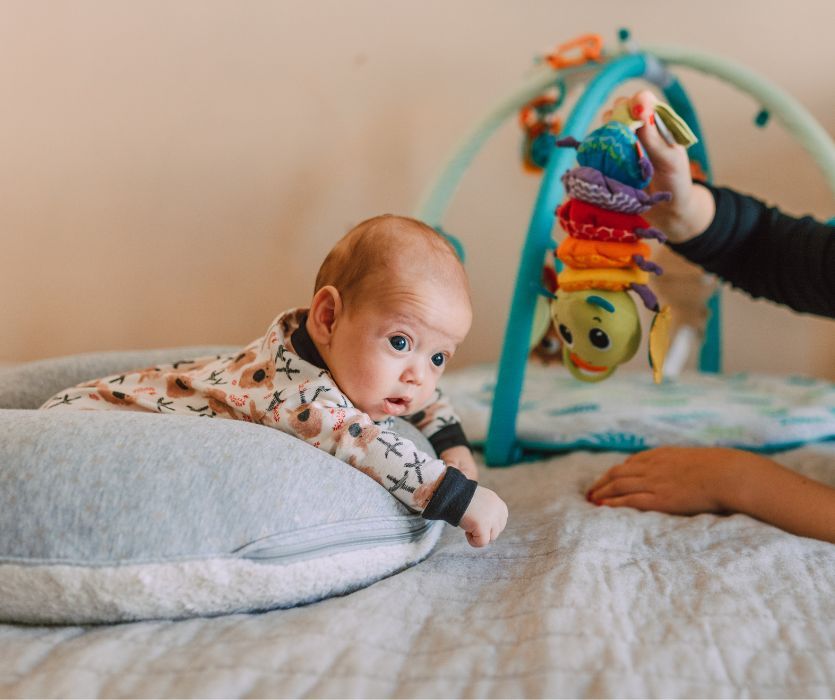 A newborn baby lies on a nursing pillow looking at a colorful caterpillar toy hanging from a play gym.