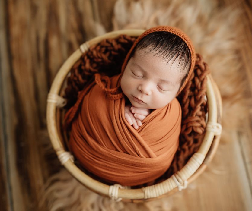 Sleeping newborn swaddled in orange fabric, tucked into a round wicker basket on a rustic wood background.