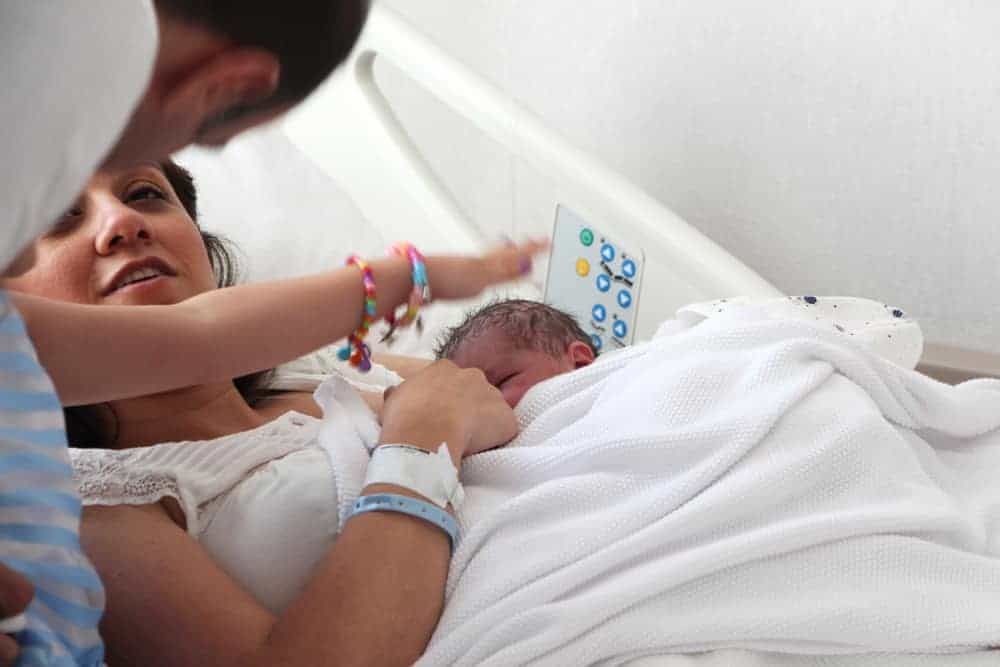 A mother in a hospital bed holds her newborn baby as a partner looks on and a child reaches toward them.