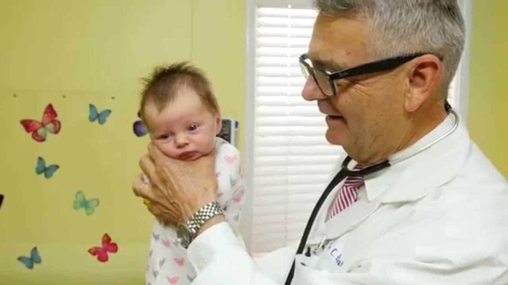 A pediatrician in a white lab coat holds a baby upright during a medical examination in a colorful office.