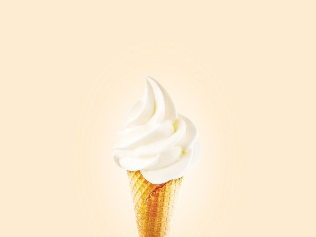 Swirl of white soft serve ice cream in a wafer cone against a cream-colored background.