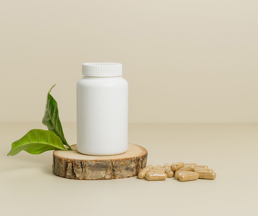 A white pill bottle on a wood slice next to a pile of placenta capsules and green leaves.