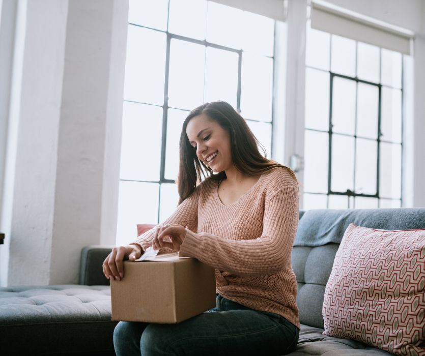 A smiling woman sits on a couch and opens a cardboard delivery box in a bright room with large windows.