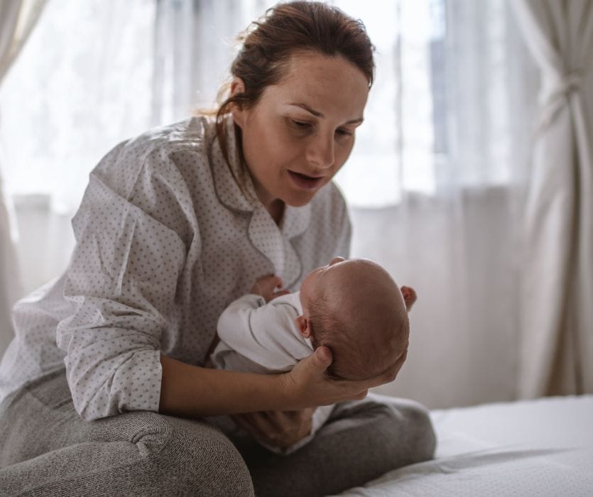 Woman in pajamas sitting on a bed and looking down anxiously while holding a newborn baby.
