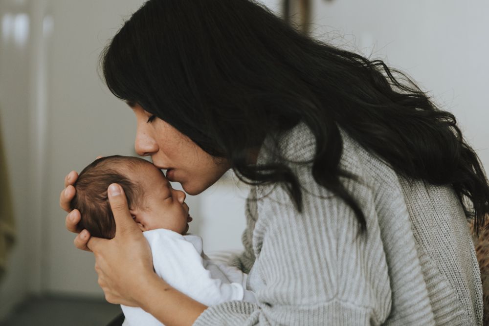 A woman with long dark hair tenderly kisses a sleeping newborn baby on the forehead while cradling them in her hands.