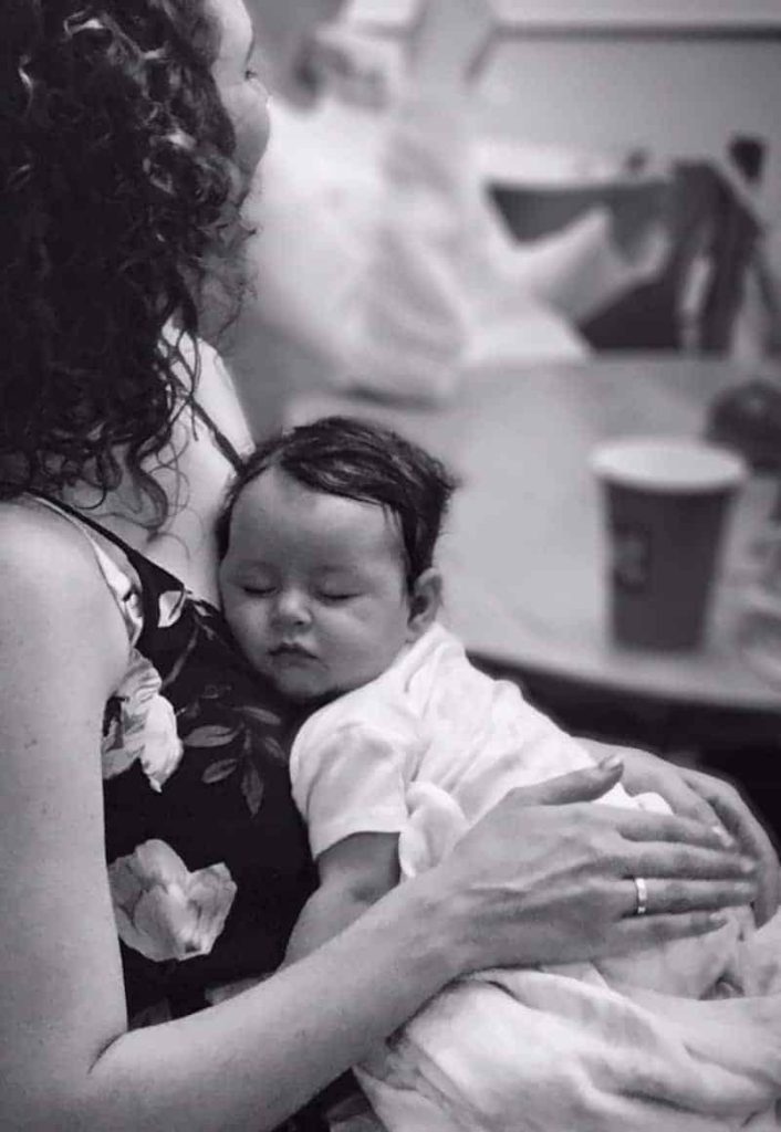Black and white photo of a woman with curly hair holding a sleeping newborn baby against her chest.