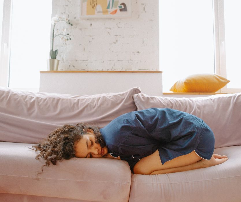 Woman in blue pajamas curled up in a fetal position on a light purple sofa, appearing to be in physical discomfort.