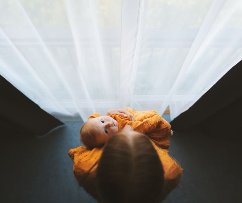Top-down view of a mother holding her infant in orange clothing against a backdrop of white curtains.