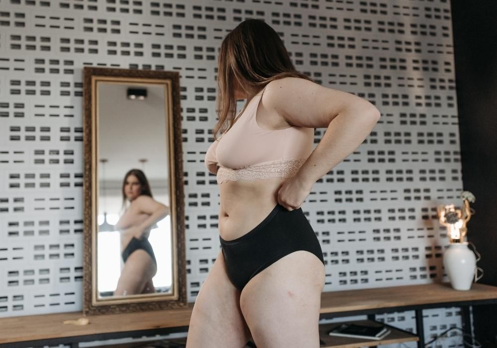 Woman wearing a peach lace bra and black high-waisted postpartum underwear standing next to a mirror.
