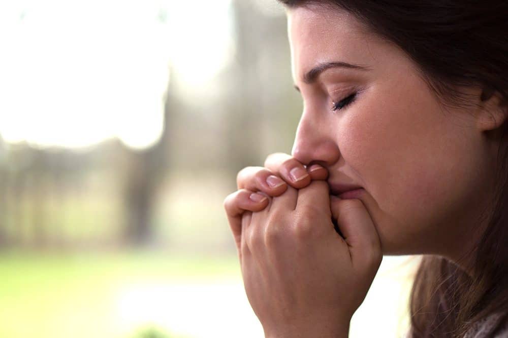 Close-up profile of a woman with her eyes closed and hands clasped to her mouth, appearing distressed or sad.