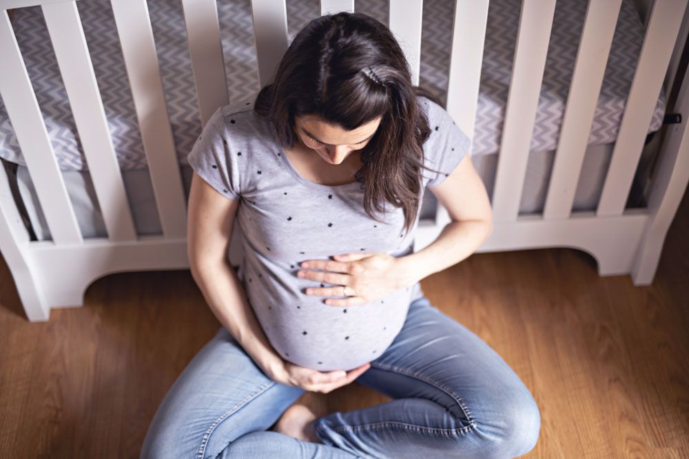 Pregnant person sitting on the floor in front of a white crib, holding their bump.