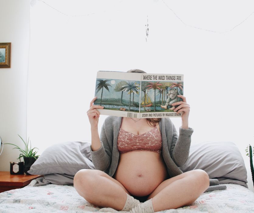 A pregnant person sits cross-legged on a bed, holding the book Where the Wild Things Are in front of their face.