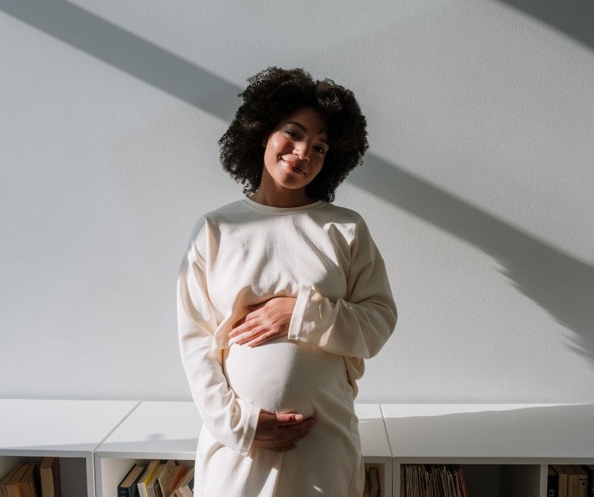 A smiling pregnant woman with curly hair stands in sunlight, holding her baby bump with both hands.