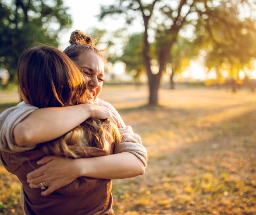 Two women sharing an emotional embrace in a sunlit park at golden hour.