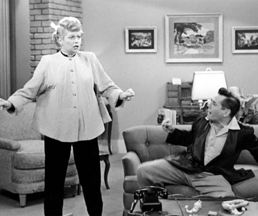 Black and white scene of Lucille Ball in a maternity smock looking surprised while Desi Arnaz reacts from a sofa.