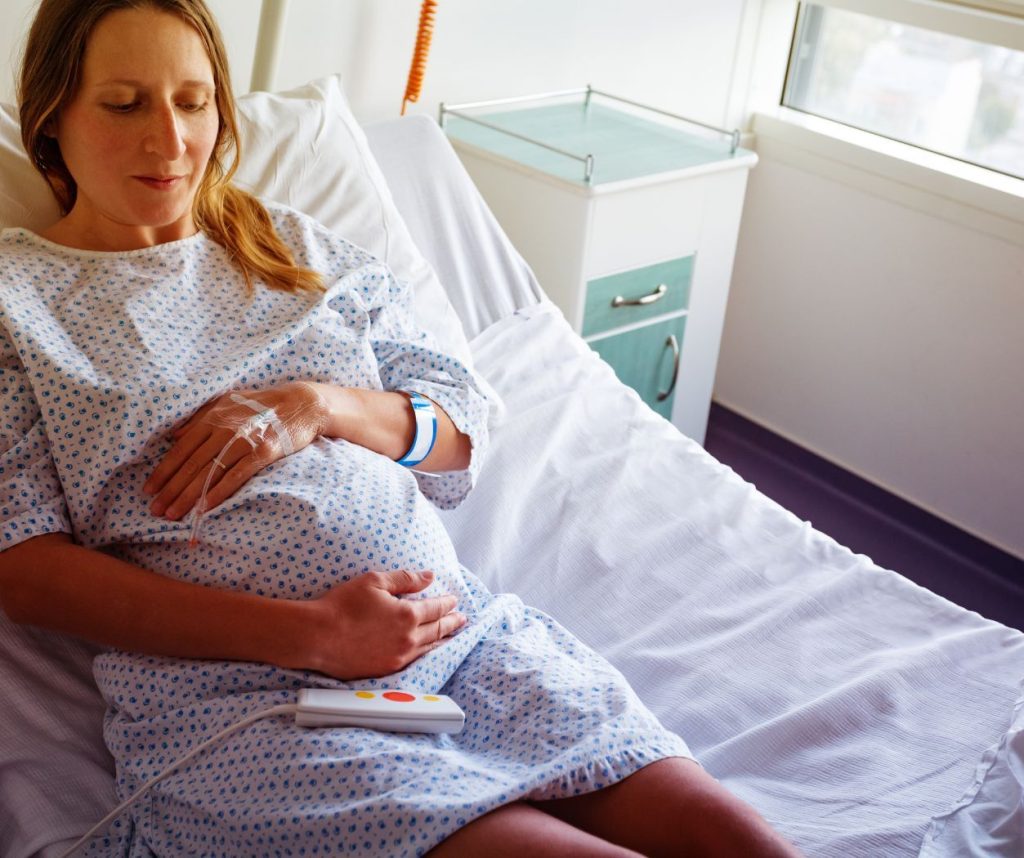 Pregnant woman in a hospital gown sitting in bed, holding her stomach with an IV in her hand during labor induction.