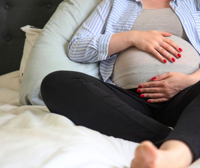 A pregnant woman sitting on a bed resting her hands on her stomach.