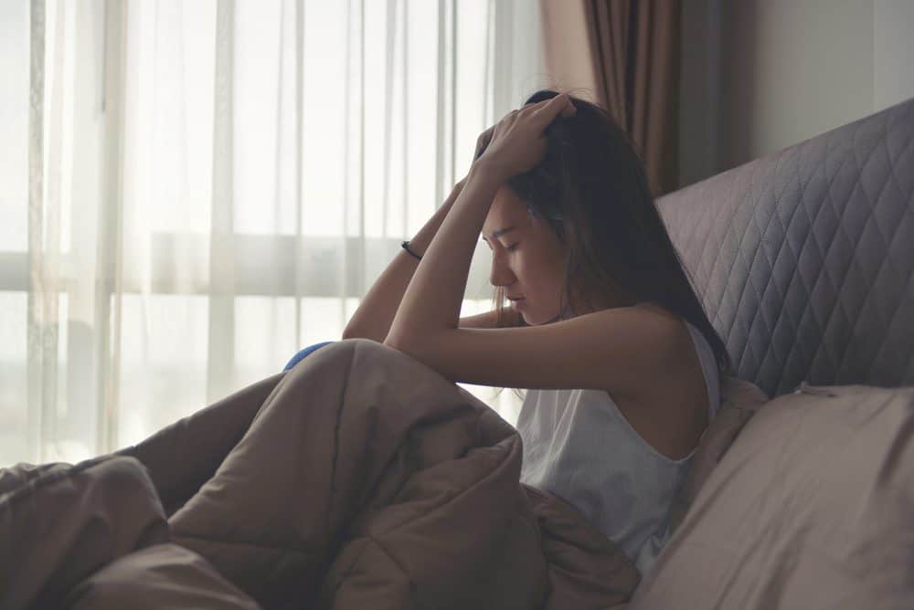 A woman sits up in bed with her hands on her head, looking down with a distressed expression.